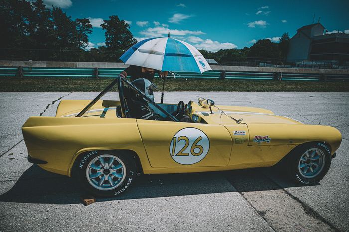The driver of this 1962 GSM Dart is shaded by an umbrella while they wait in the false grid for their turn on the track.